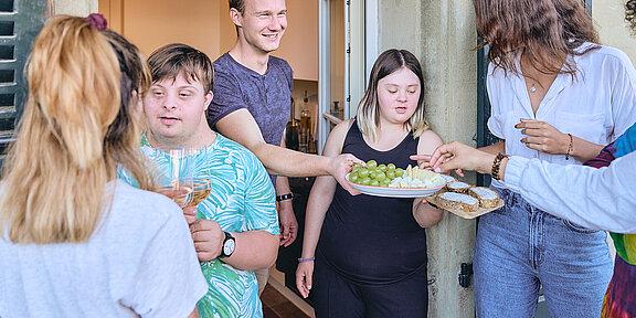 Eine Gruppe junger Menschen mit und ohne Beeinträchtigung, die auf einem Balkon stehen. Zwei Personen servieren Apéro.